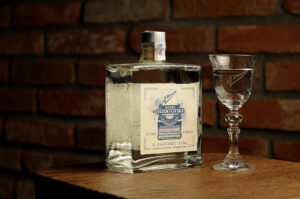 Wódka z wódką żytnią Redaktorską. Butelka kwadratowa z ładną etykietą. Obok stoi kieliszek z wódką. W tle ściana.