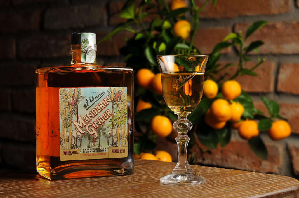Duża butelka likieru Mandarin Ginger w kwadratowej butelce z kolorową etykietą. Obok stoi pełnu kieliszek. W tle drzewko mandarynkowe.