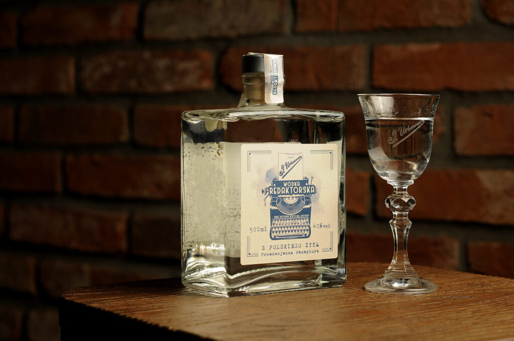 Wódka z wódką żytnią Redaktorską. Butelka kwadratowa z ładną etykietą. Obok stoi kieliszek z wódką. W tle ściana.
